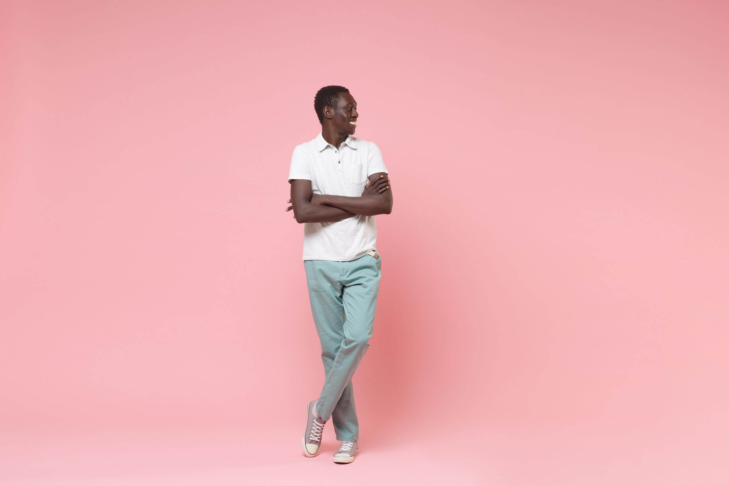 Smiling young african american man in white polo shirt, turquoise trousers posing isolated on pastel pink background. People lifestyle concept. Mock up copy space. Holding hands crossed looking aside.